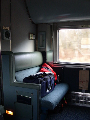 [A double-wide padded seat up against a wall with a large window beside it. A duffle bag and a coat sit on the seat.]